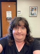 Heather is taking a selfie and has long brown curly hair and is wearing a blie v neck t shirt.